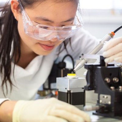 a woman in safety glasses looks closely at a yellow disk on a stand